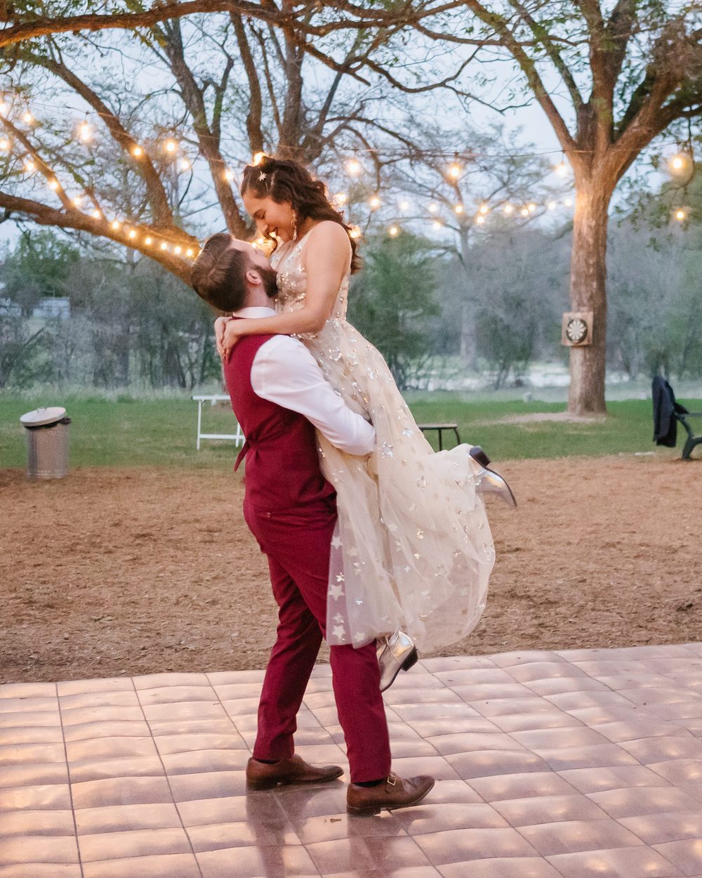 Groom lifting his bride high during their first dance under string lights