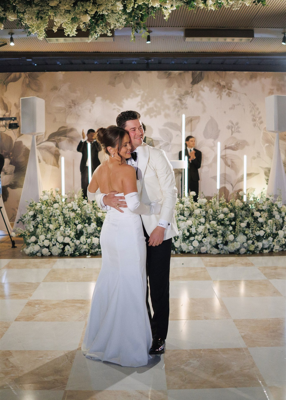 Bride and groom in a close embrace on a marble checkered floor surrounded by white flowers