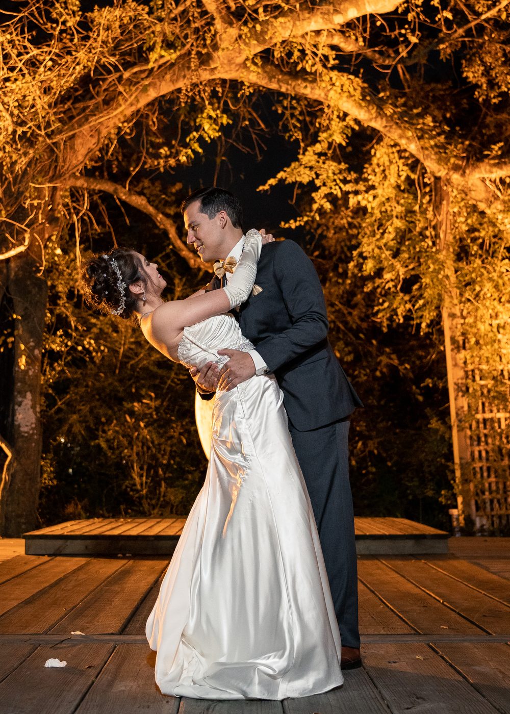 Groom dipping his bride under a glowing golden tree canopy at night